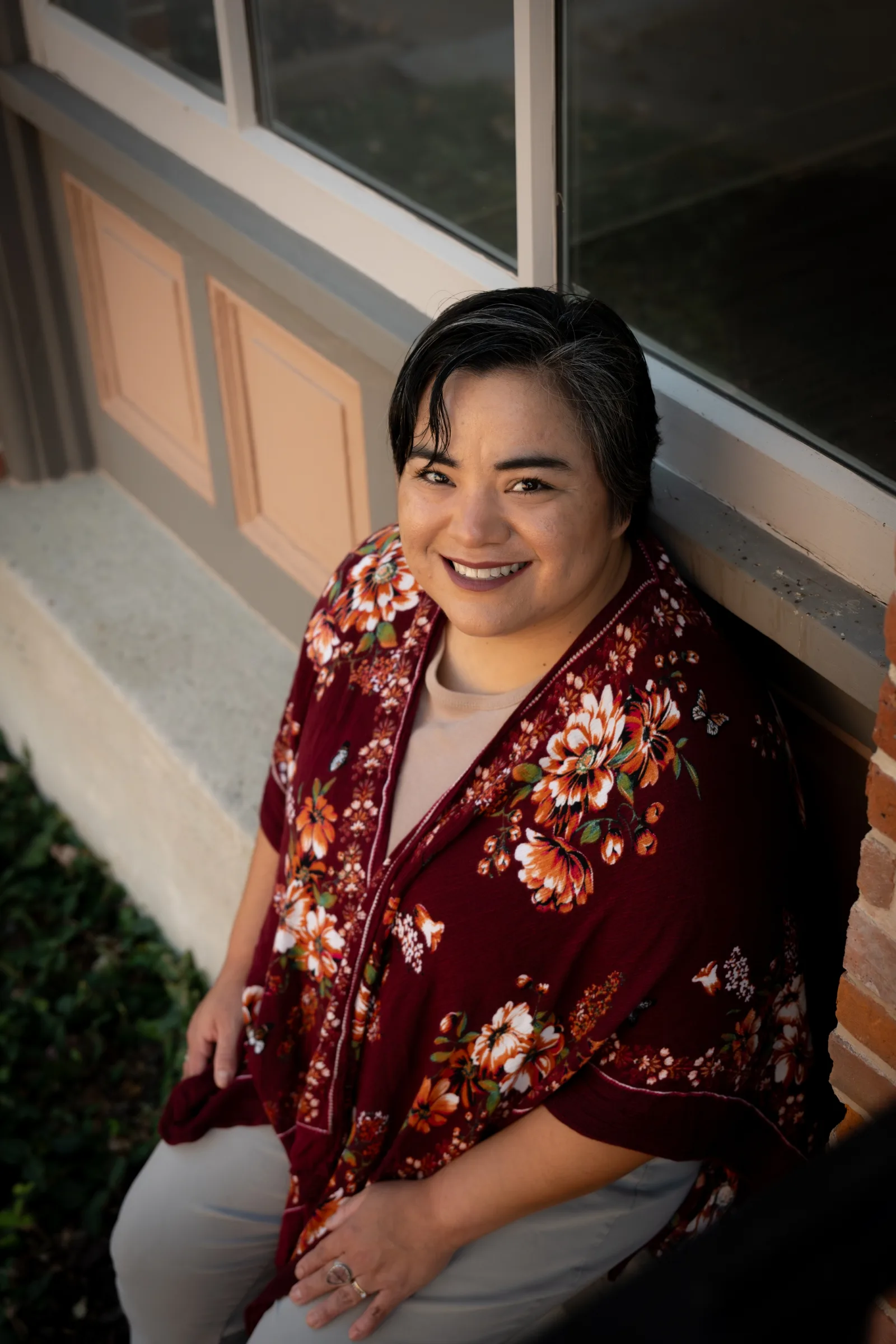 Portrait of therapist Alyssa sitting outdoors by a window, smiling and wearing a floral top