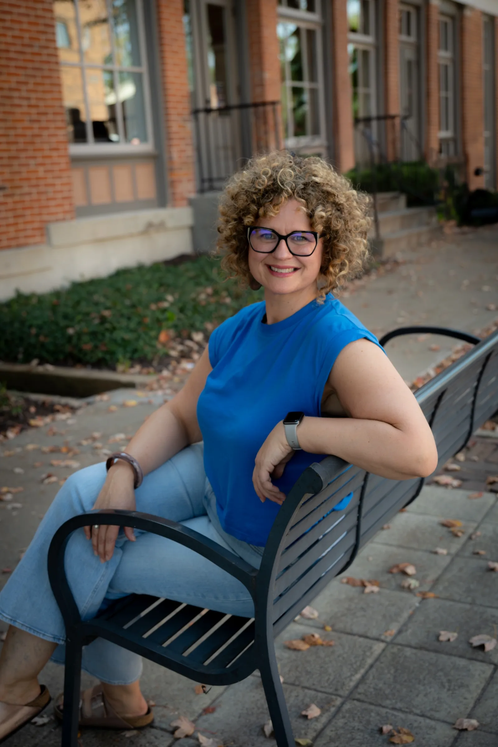 Portrait of therapist Amanda sitting on an outdoor bench, smiling with curly hair and glasses.