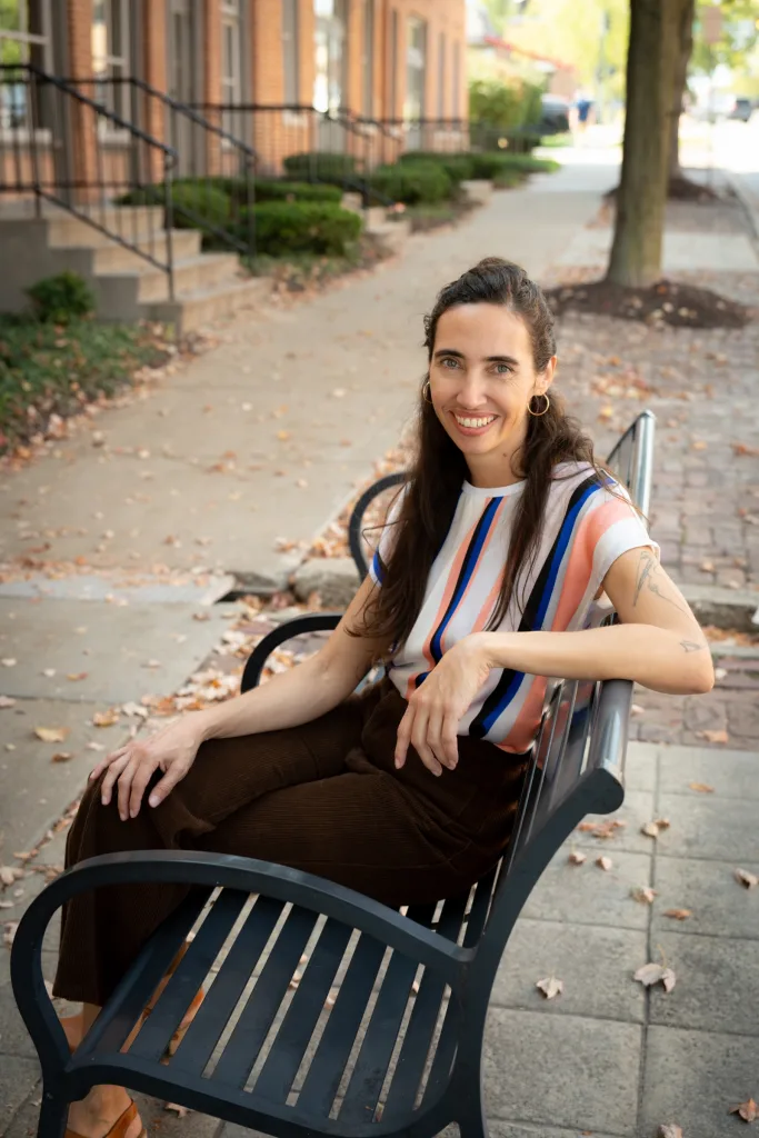 Portrait of therapist Candida sitting on an outdoor bench, smiling and wearing a striped blouse.