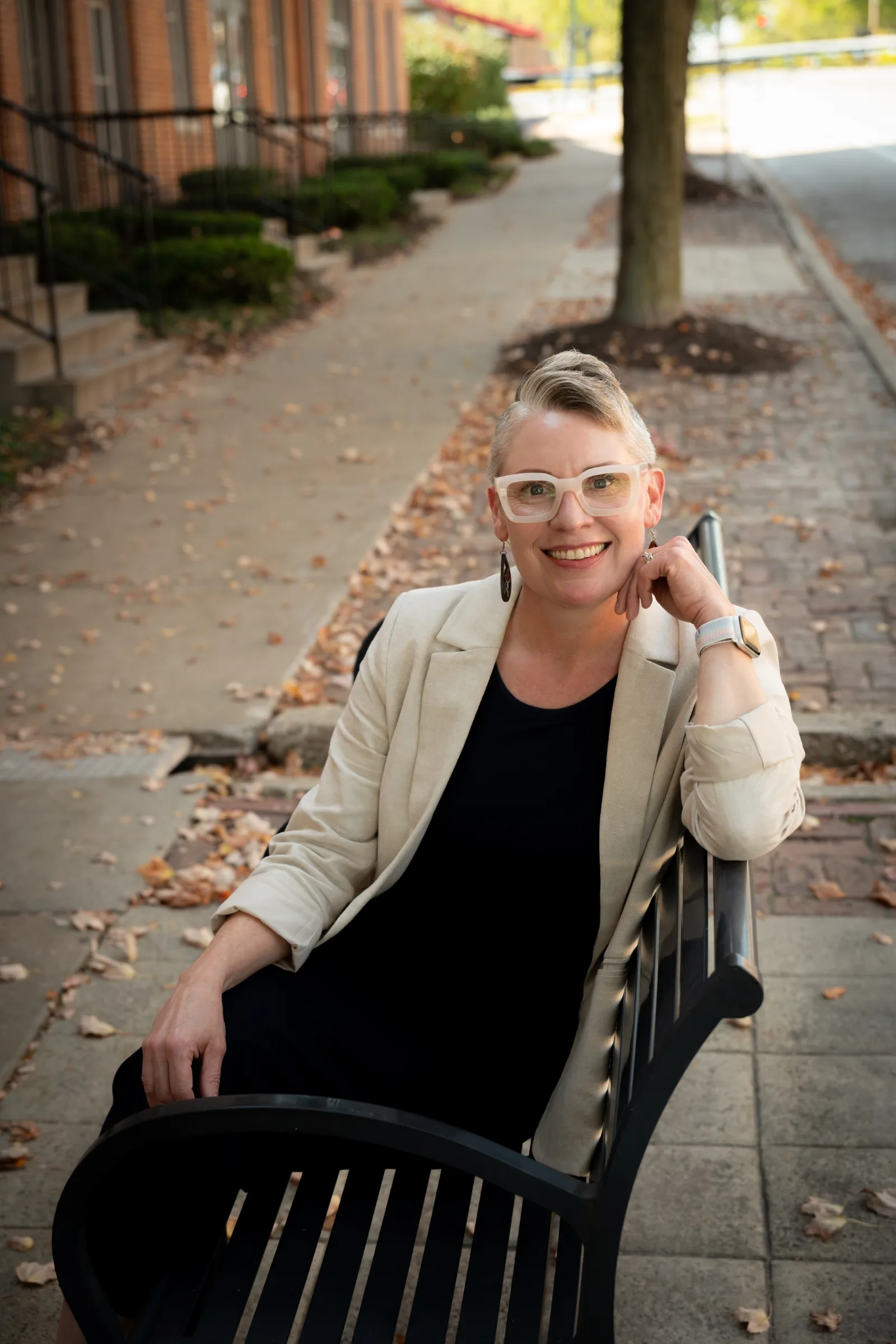 Portrait of therapist Darcie sitting on an outdoor bench, smiling and wearing a light blazer and glasses.