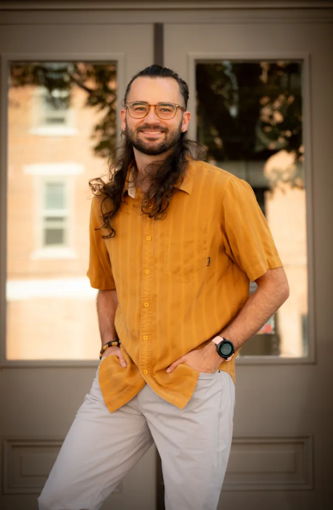 Portrait of therapist Miles standing outdoors in front of a doorway, smiling with hands in pockets.